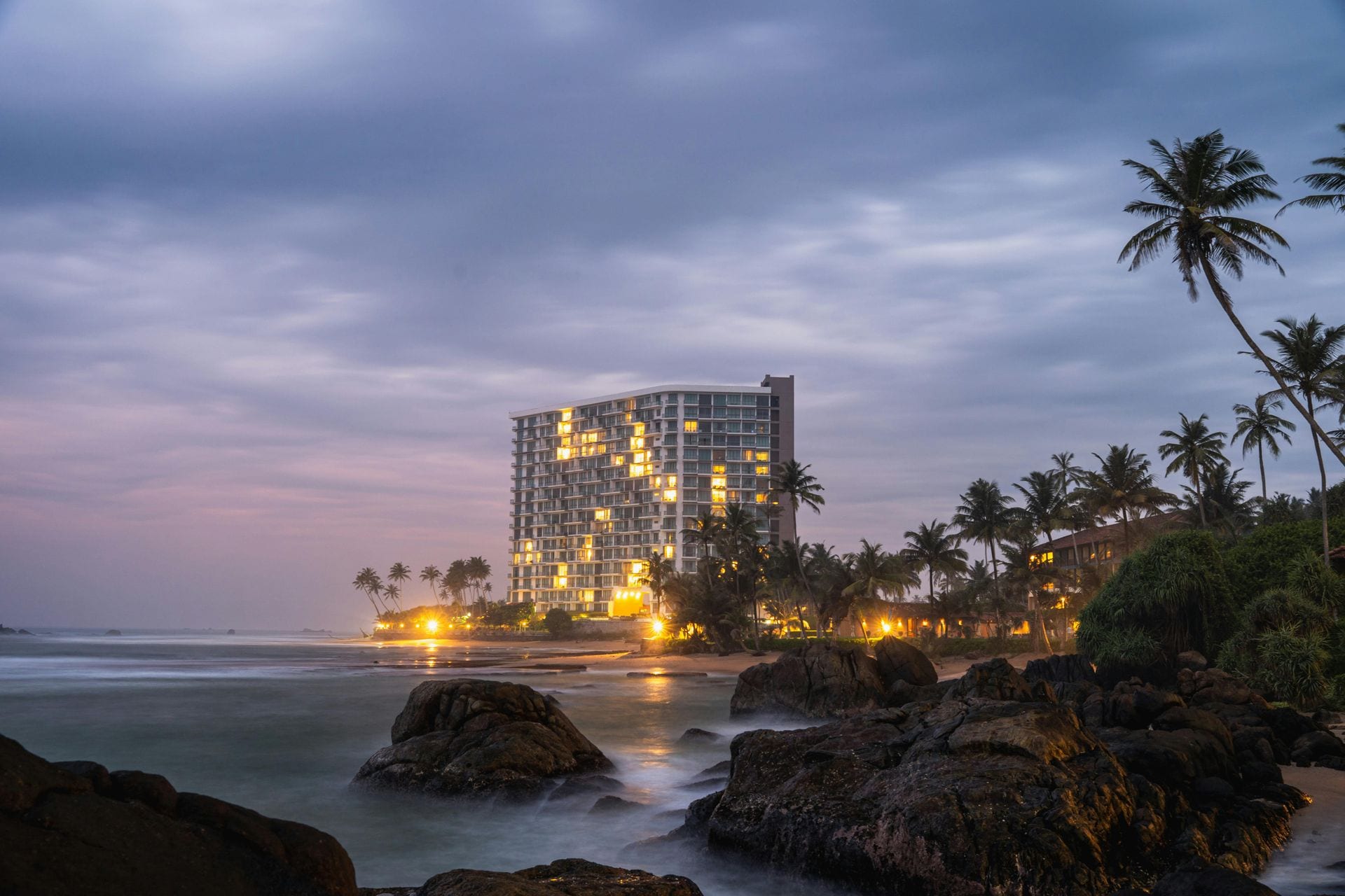 Hotel em frente ao mar ao crepúsculo, rochas em primeiro plano — distribuição costeira