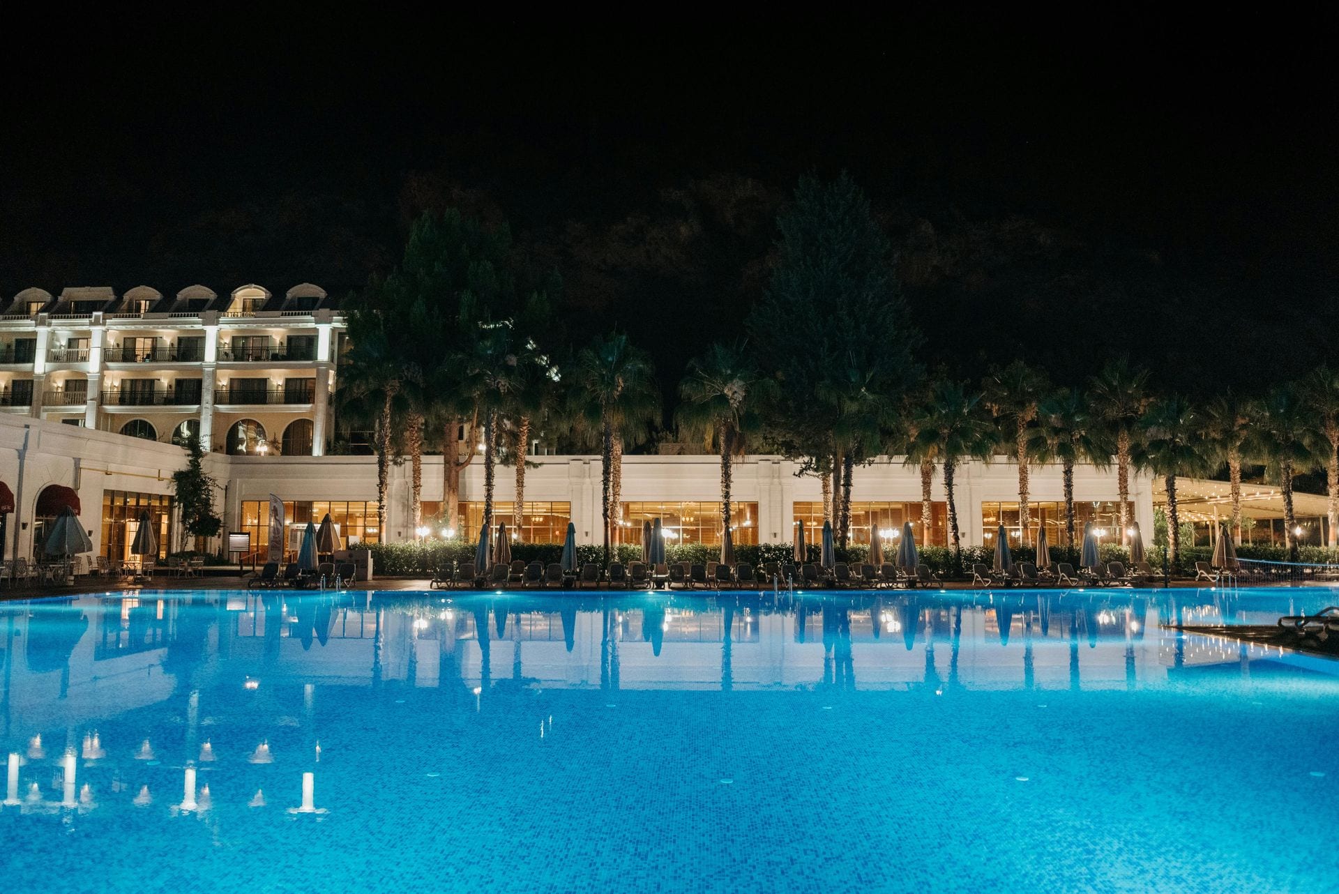 Hotel iluminado ao anoitecer refletido em grande piscina entre palmeiras — presença