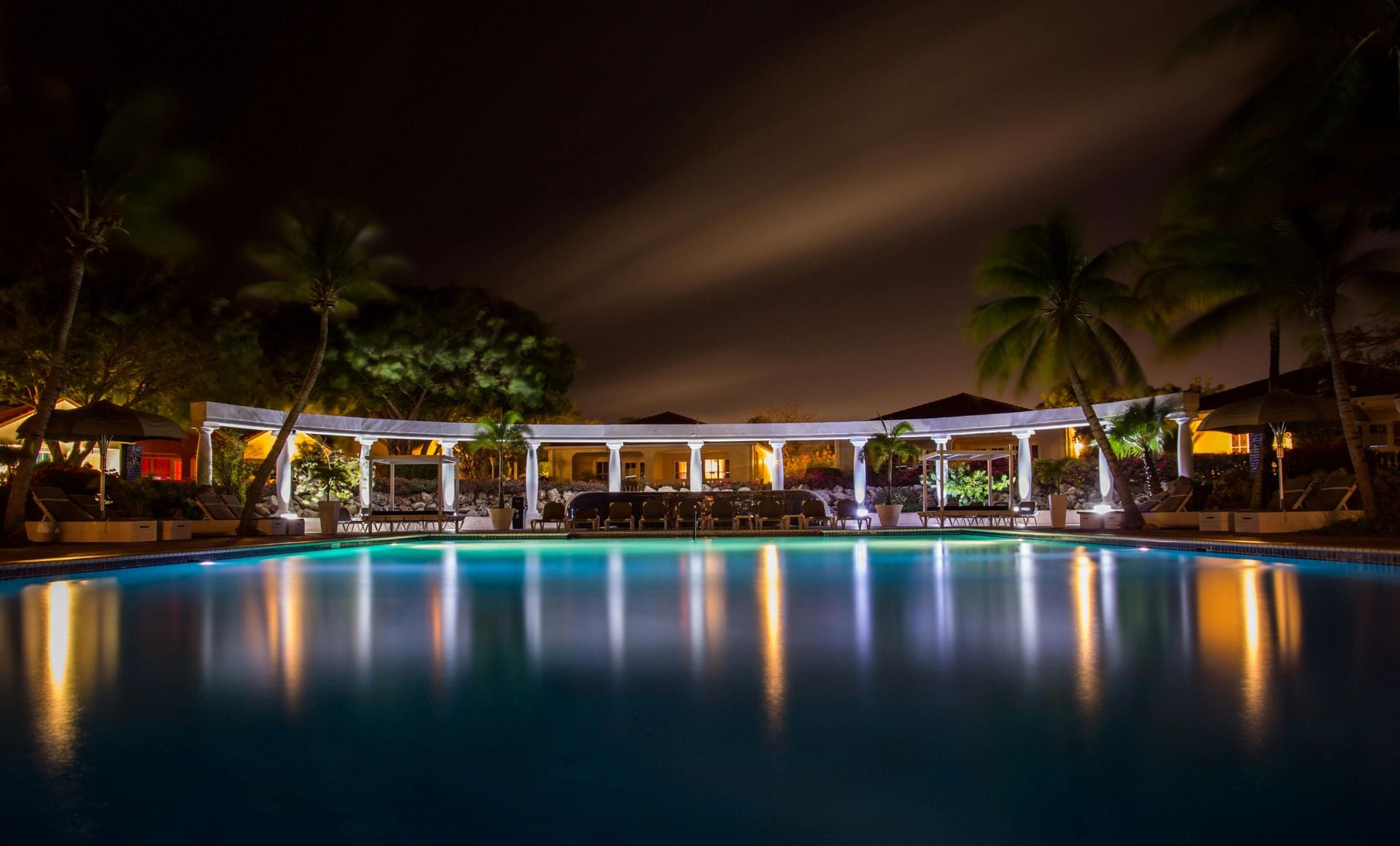 Piscina iluminada à noite com palmeiras e edifício em tom amarelo — momento de pausa