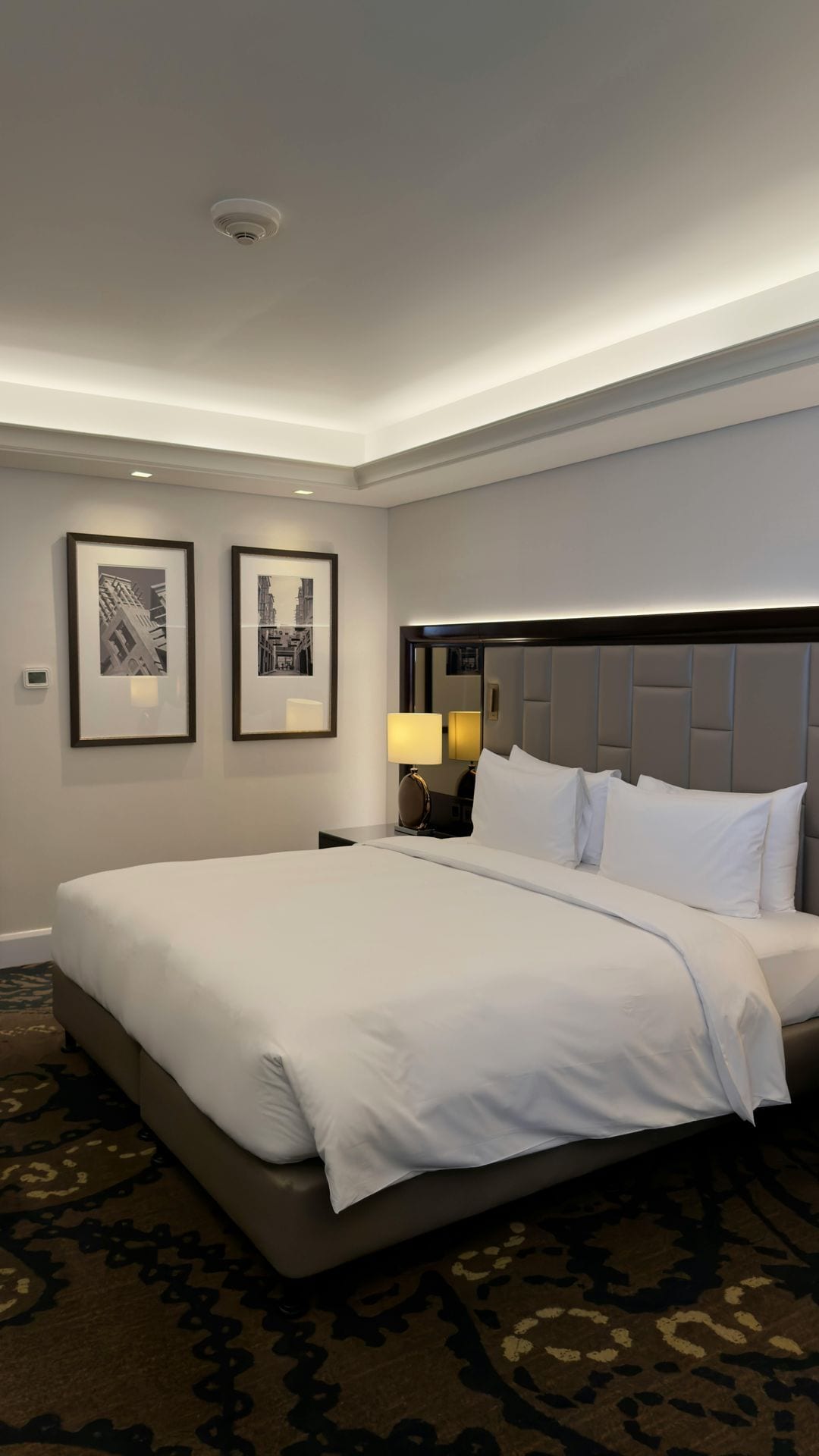 Quarto de hotel clássico em tons quentes com cabeceira capitonê cinza e fotografias emolduradas